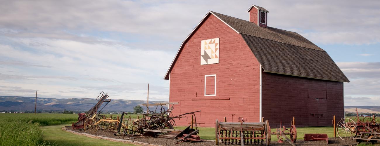 Red hey barn surrounded by early 1900 farm equipment. Green hills in the distance and light grey clouds against blue sky. 