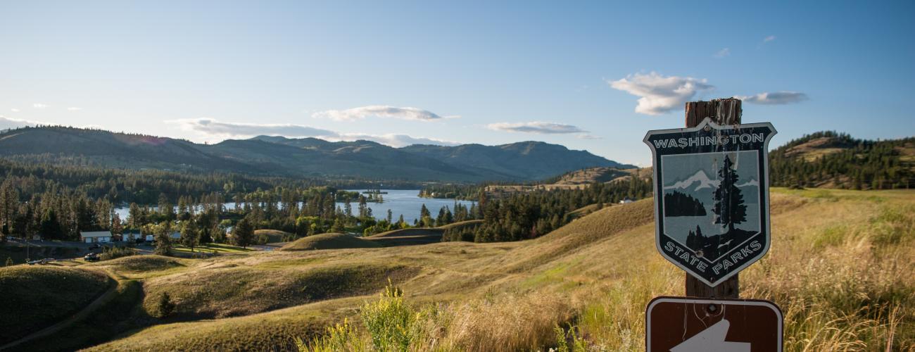 A Washington State Parks logo sign points towards Lake Curlew that is visible beyond golde, grassy, rolling hills.