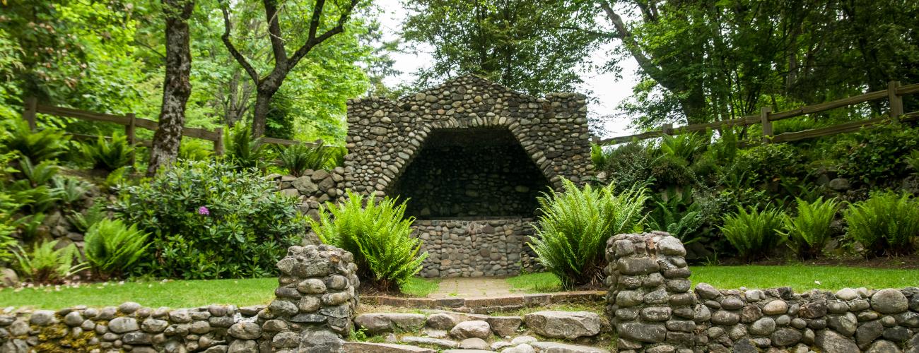 The grotto is made up of hundreds of stones of varying colors surrounded by multiple bushes and lush green trees. There is a path leading up to the grotto with a few steps. 