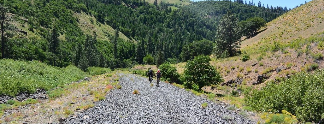 Two cyclists ride on a wide gravel trail with tree covered hillsides on either side.