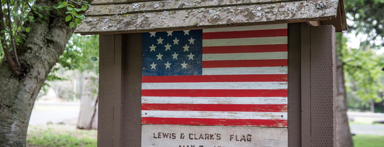 Brown roofed sign with a painted replica of Lewis and Clark's flag from 1806 with 15 stars on a blue background and 15 red and white stripes