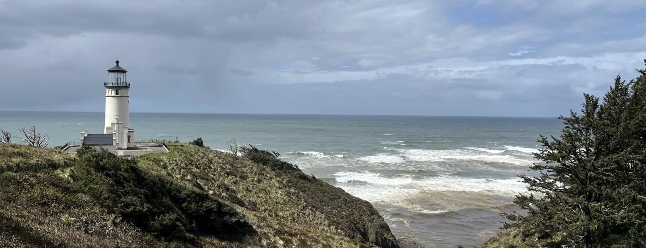 Standing on a rocky shore, looking towards the ocean, North Head Lighthouse is white against a stormy grey sky.