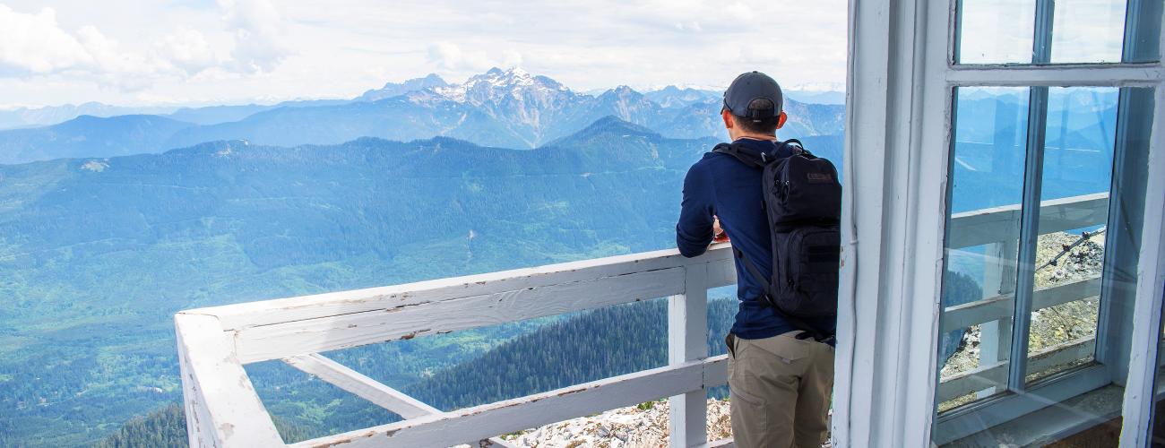 A hiker takes a break while enjoying the view of forested mountains and a snowcapped mountain with a cloudy sky from the lookout tower's deck, with white chipping paint. 