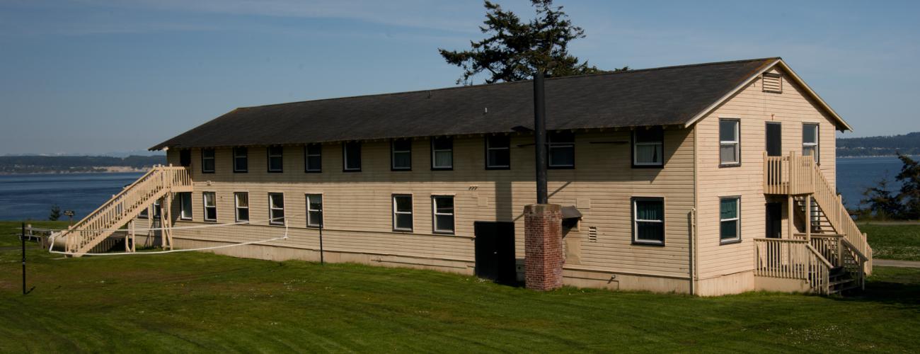 A two-story tan dorm building sits on a lush, green lawn with an evergreen tree standing behind the building, the Puget Sounds is seen to the left of the building. On the grass is a volleyball net. 