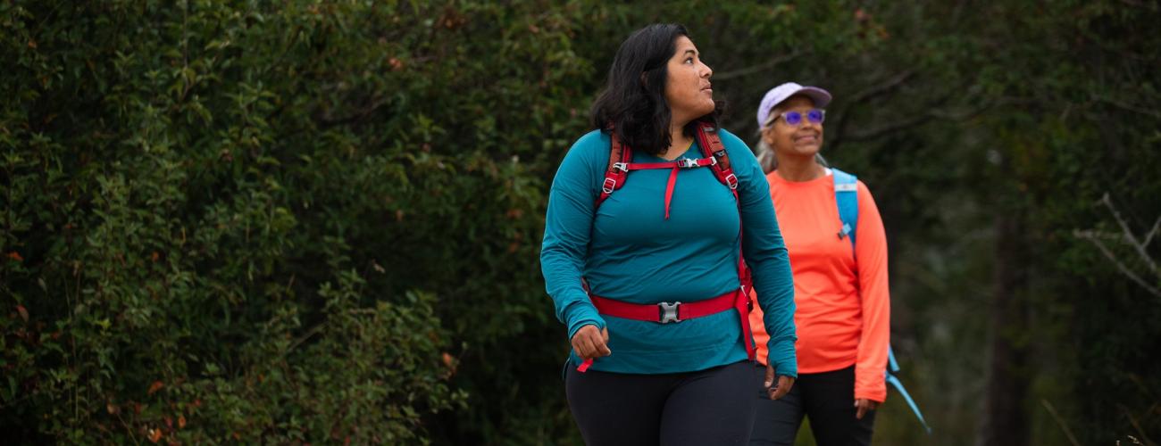 Two women, one in blue and one in orange, hike in the forest wearing backpacks.