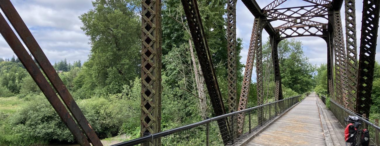 A view looking down an old railroad bridge made of iron on the Willapa Hills State Park Trail. The rails have been replaced with boards and a bicycle is leaning on the rail on the  right side. To the left the Willappa River is seen flowing under the bridge and the  blue sky above is filled with fluffy white clouds.