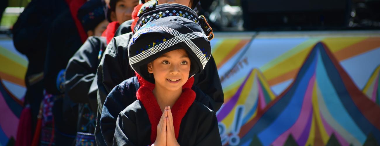 A group of young Mien dancers dress in traditional clothing. The girl in the front smiles and holds up her hands in praying gesture. 