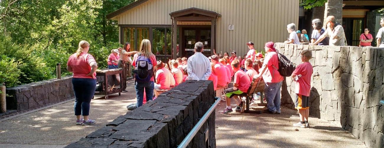 A large group of children sit outside a building and listen while a park ranger speaks.