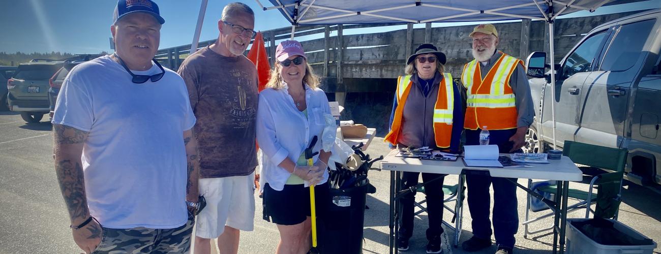 A group of five volunteers with litter pickers stand with smiles under a blue canopy. A wooden pier sits in the background.