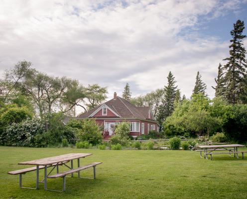 Red house built in 1908 hidden in the middle of pine trees, lilac and rose bushes. Manicured lawn with picnic tables. Large white and grey clouds in the sky with blue sky coming through.