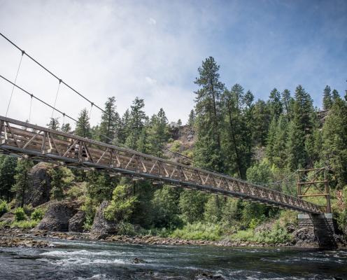 A suspension bridge spans the Spokane River with trees covering the far bank and partly cloudy skies.