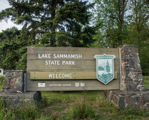 Entrance sign at Lake Sammamish State Park. Features the pre-2023 logo, the name of the park, the words "welcome" and "cooperating agencies" with accompanying identifiers for the cooperating agencies. These small signs are however unreadable. In the background lush, green leaved trees, evergreens, and grass are visible. 