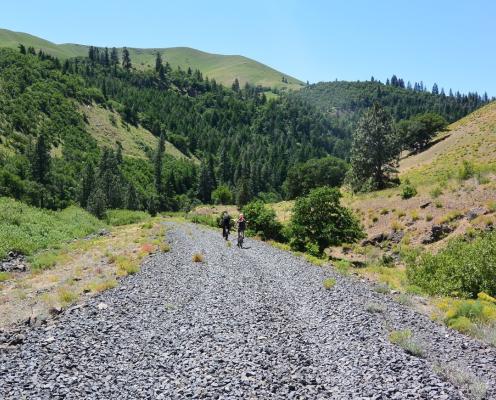 Two cyclists ride on a wide gravel trail with tree covered hillsides on either side.