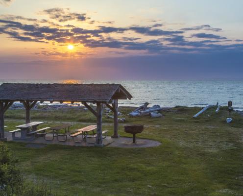 A wooden picnic shelter with picnic tables, surrounded by green grass, sits near the beach with driftwood nearby. The waters of the Strait of Juan de Fuca with a blue, yellow and pink sky as the sun sets behind the shelter.