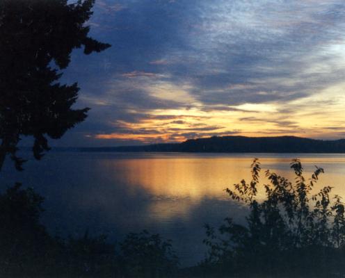 Dark shadows of trees and bushes frame an orange and dark gray late sunset reflected in the placid waters fronting Triton Cove State Park.