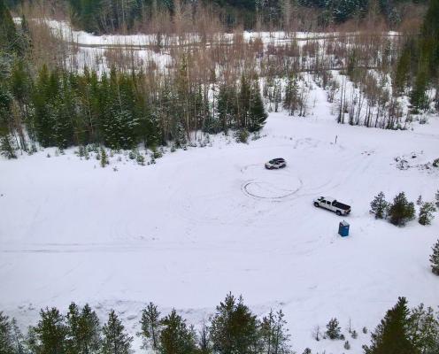 A large unplowed parking area bordered by young trees, with a narrow plowed road in the distance. A porta-potty is present.