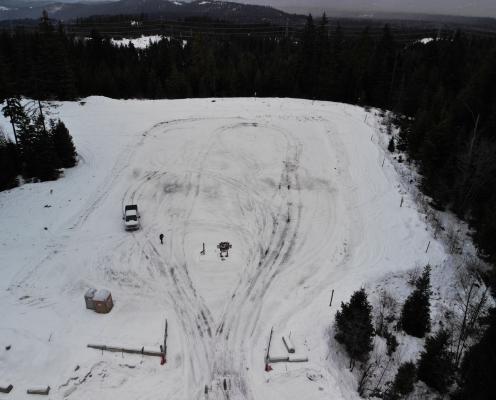 A rectangular parking area covered with a layer of thin snow and tire tracks. Two porta-potties sit in the lower left corner.