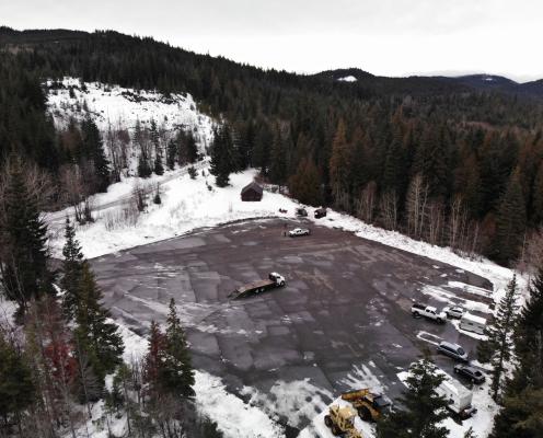 Large square paved parking lot plowed clean surrouned by pine trees next to a forest road with light snow. A warming hut and pit toilet hut stand in the upper right corner and several trucks are parked along the right edge of the lot.