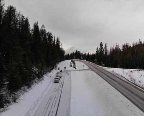 A plowed highway turns onto a plowed forest road covered by a thin layer of snow. A truck with a trailer and a porta-potty stand next to an information board.