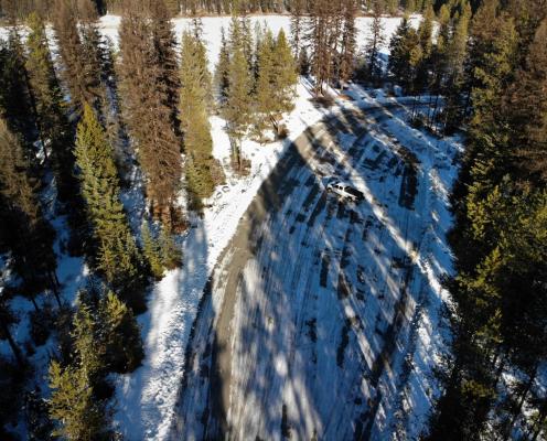 Overhead view of a plowed parking area covered by strips of thin snow and surrounded by pine trees.