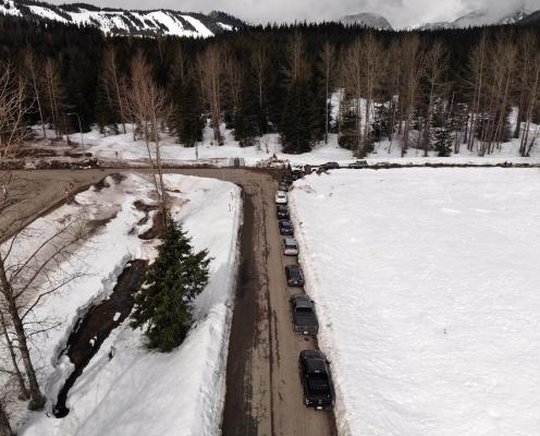 Paved forest road next to a highway, both plowed clean and bordered by thick unplowed snowy ground. A line of trucks and three porta-potties lie along the edge of the road.