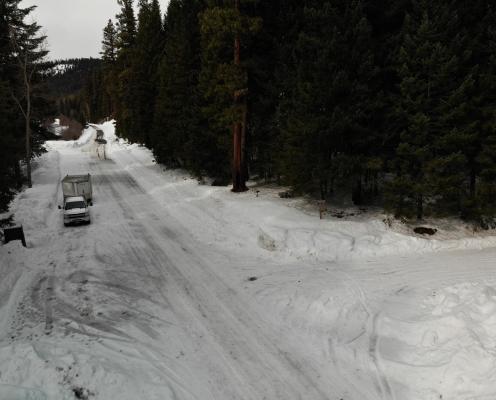 T-intersection of two forest roads covered thinly with snow surrounded by pine trees. A porta-potty sits in the corner half-buried in snow.