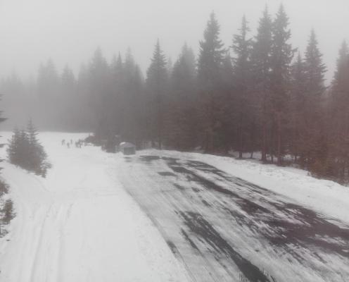 Foggy view of an icy parking area surrounded by thick snow-covered ground. A pit toilet hut and several skiiers stand on one end of the lot.