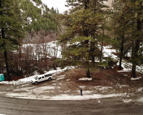 A plowed pull-off parking area with patchy snow next to a forest road. A porta-potty sits on the left side.