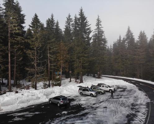 A plowed highway curves around a plowed parking area next to a forested trailhead.