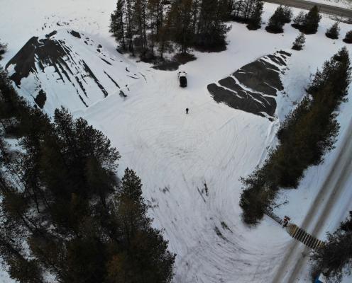 Parking area with compacted snow, flanked by two snow-covered pyramid-shaped hills and scattered trees along the edges. A plowed road appears in the corner.