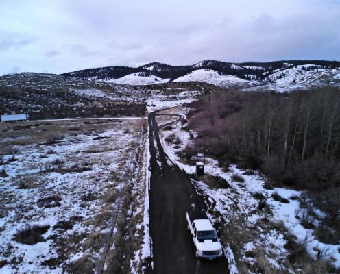 A plowed highway runs between a forest and a meadow with patchy snow with snowy hills in the background.