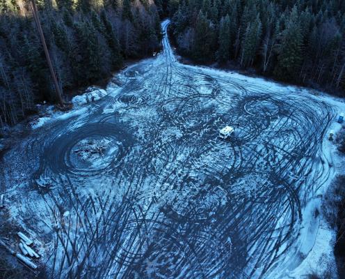 A large parking area with thin layer of ice and snow overrun by numerous tire tracks, surrounded by a thick dense forrest. A porta-potty sits in the corner.