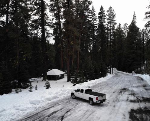 Icy forest road opening onto a wider ice-covered parking area bordered by unplowed snow and snowy ground. A picnic shelter sits a 50 feet back from the edge of the lot.