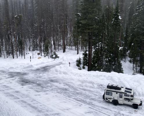 A forest road leading to a parking area, both covered in a thin layer of snow and ice, next to a snow-covered forest. 