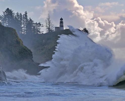 High king tide wave crashes against rocky cliffside with a lighthouse