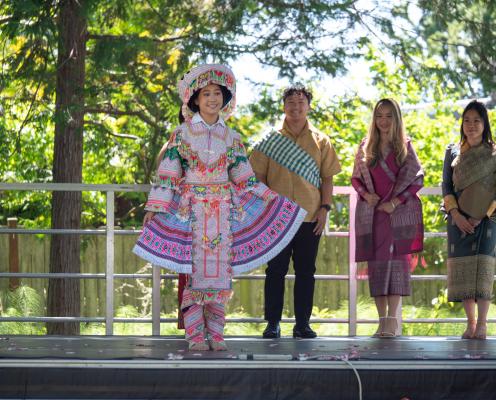 A female dresses in traditional clothing of Laos standing on stage for a fashion show. Three people dress in traditional clothing standing behind her. 