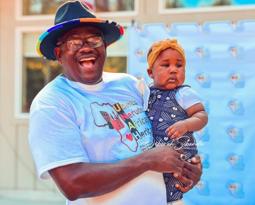 A smiling man in a hat holds a baby dressed in traditional African clothing.