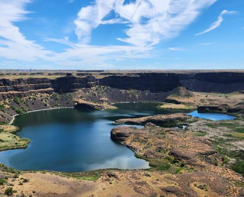 A beautiful view from the Dry Falls Visitor Center at Sun-Lakes Dry Falls State Park of the rock formations made by ice age floods. It is a sunny, blue-sky day and the land formations are brown surrounded by dry grasses and vegetation.