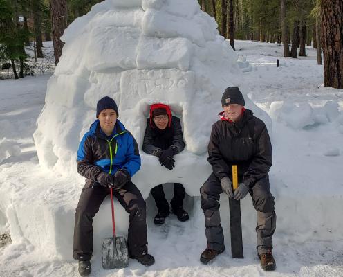 Three boys sit in front of an igloo in the snow.