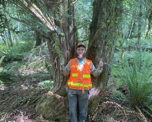 A person with a beard and work vest stands in front of a yew tree