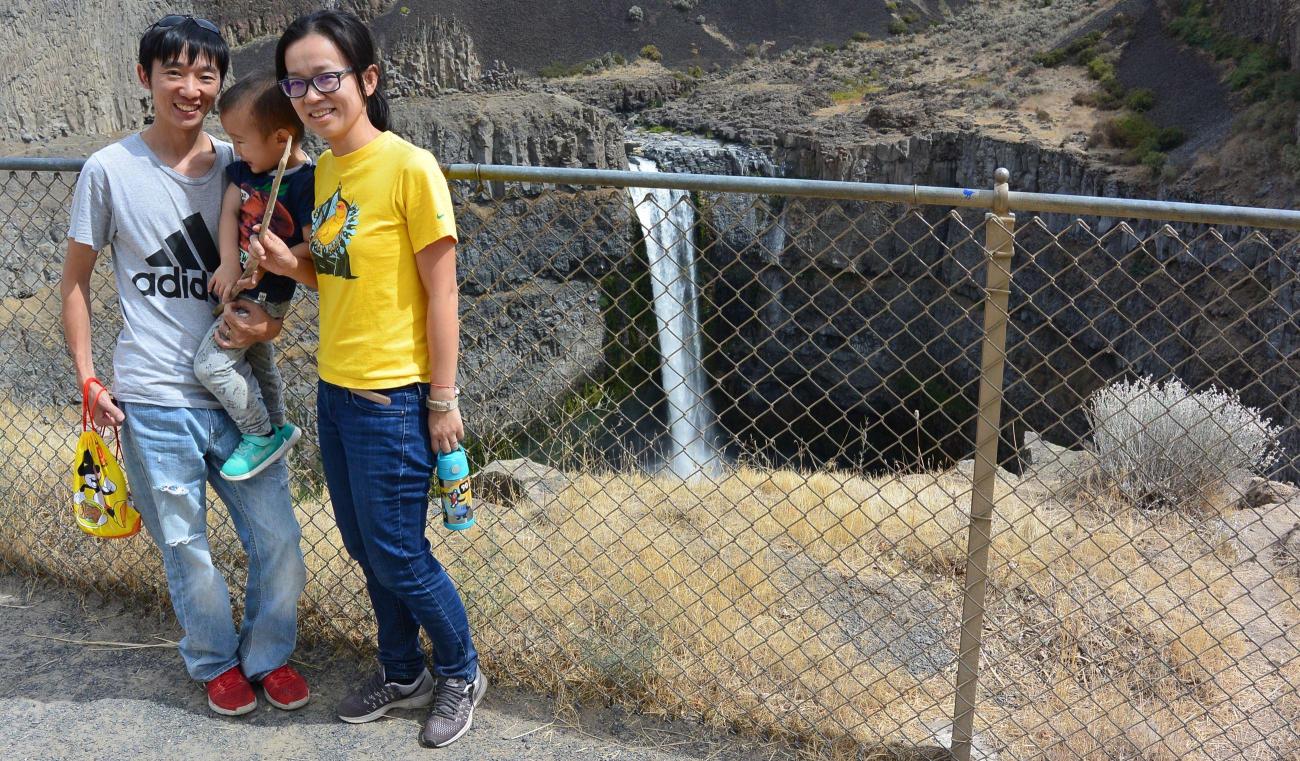 A mother and father hold a small child standing in front of Palouse Falls viewpoint behind a fence