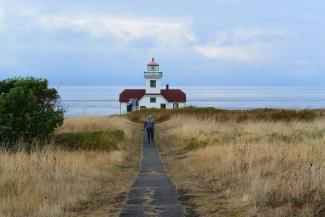 A long distance shot of Patos Island Lighthouse, a classic red and white lighthouse at the end of a long path once used to bring oil and kerosene to the lighthouse. A woman in a sweatshirt is walking toward the camera. Light brown shrubs line either side of the cement pat and there light clouds in the blue sky beyond the lighthouse.