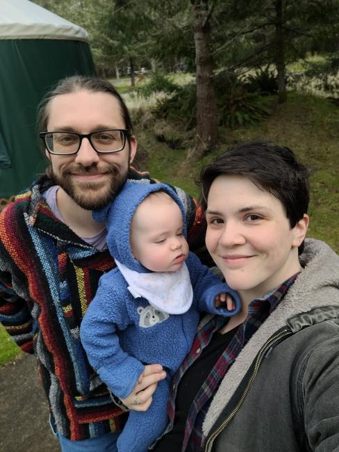 A couple and there baby smile as they stand in the forest near a yurt