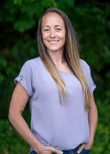 Washington State Parks Executive Assistant Becki Ellison poses in front of evergreens smiling and wearing a lavender shirt.