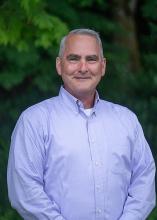 Mike Sternbeck, Washington State Parks Deputy Director poses in front of evergreen trees. He has gray hair and is wearing a lavender shirt. 