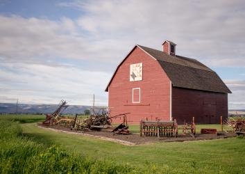 Red hey barn surrounded by early 1900 farm equipment. Green hills in the distance and light grey clouds against blue sky. 