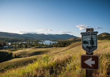 A Washington State Parks logo sign points towards Lake Curlew that is visible beyond golde, grassy, rolling hills.