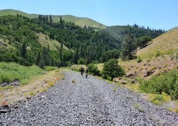 Two cyclists ride on a wide gravel trail with tree covered hillsides on either side.