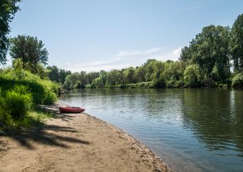 A red kayak sits on the sandy beach next to a river with small ripples reflecting the green trees and blue sky. Tall, green grass lines the edge of the sandy beach. 