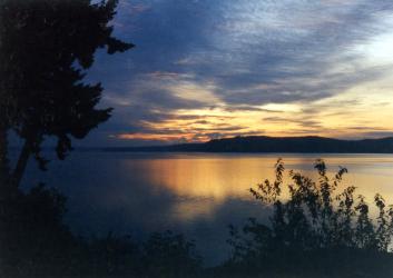 Dark shadows of trees and bushes frame an orange and dark gray late sunset reflected in the placid waters fronting Triton Cove State Park.